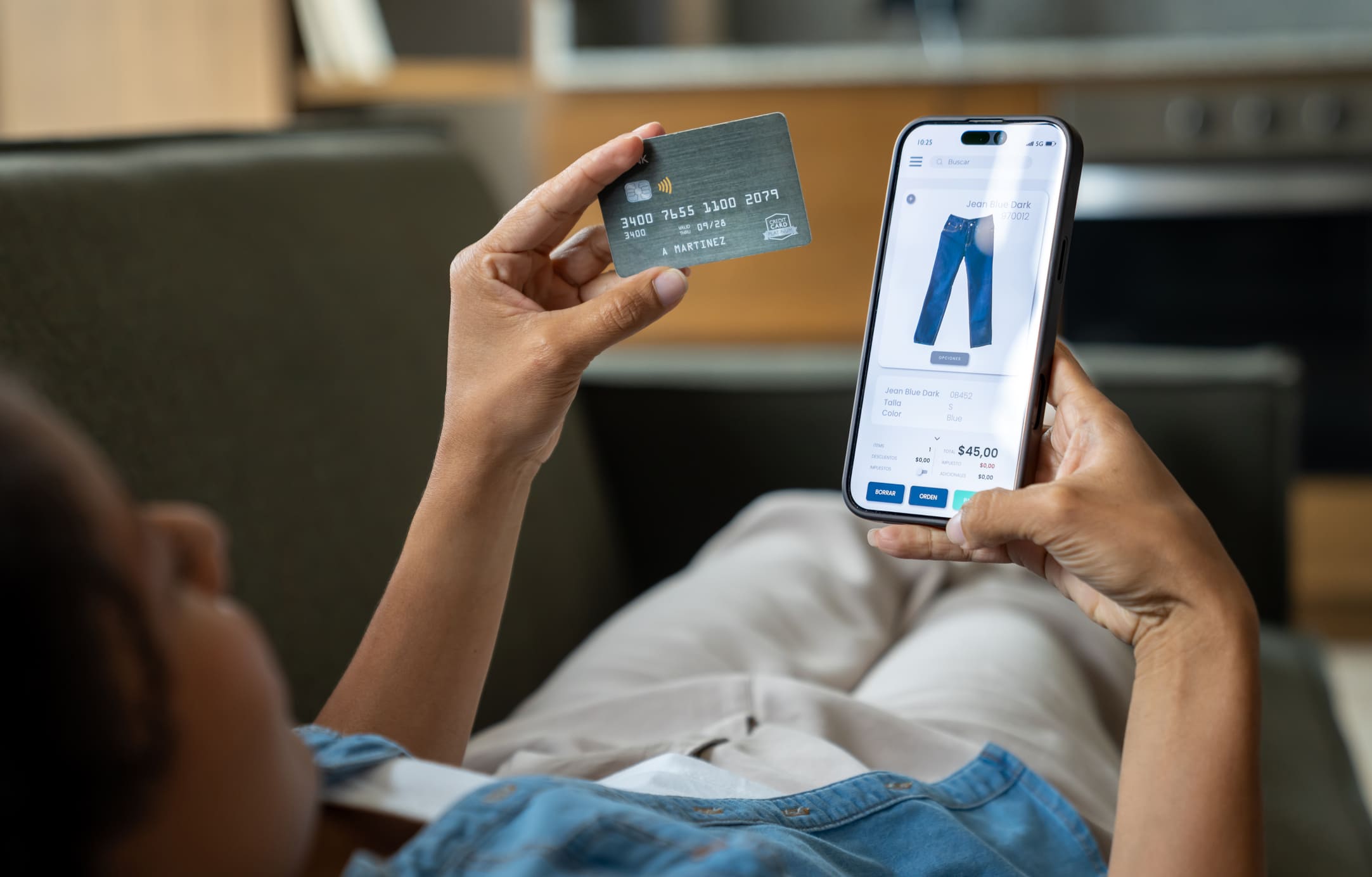 Une personne confortablement installée sur son canapé effectue un achat de vêtement sur son smartphone avec une carte bancaire, illustrant l'intégration mobile.